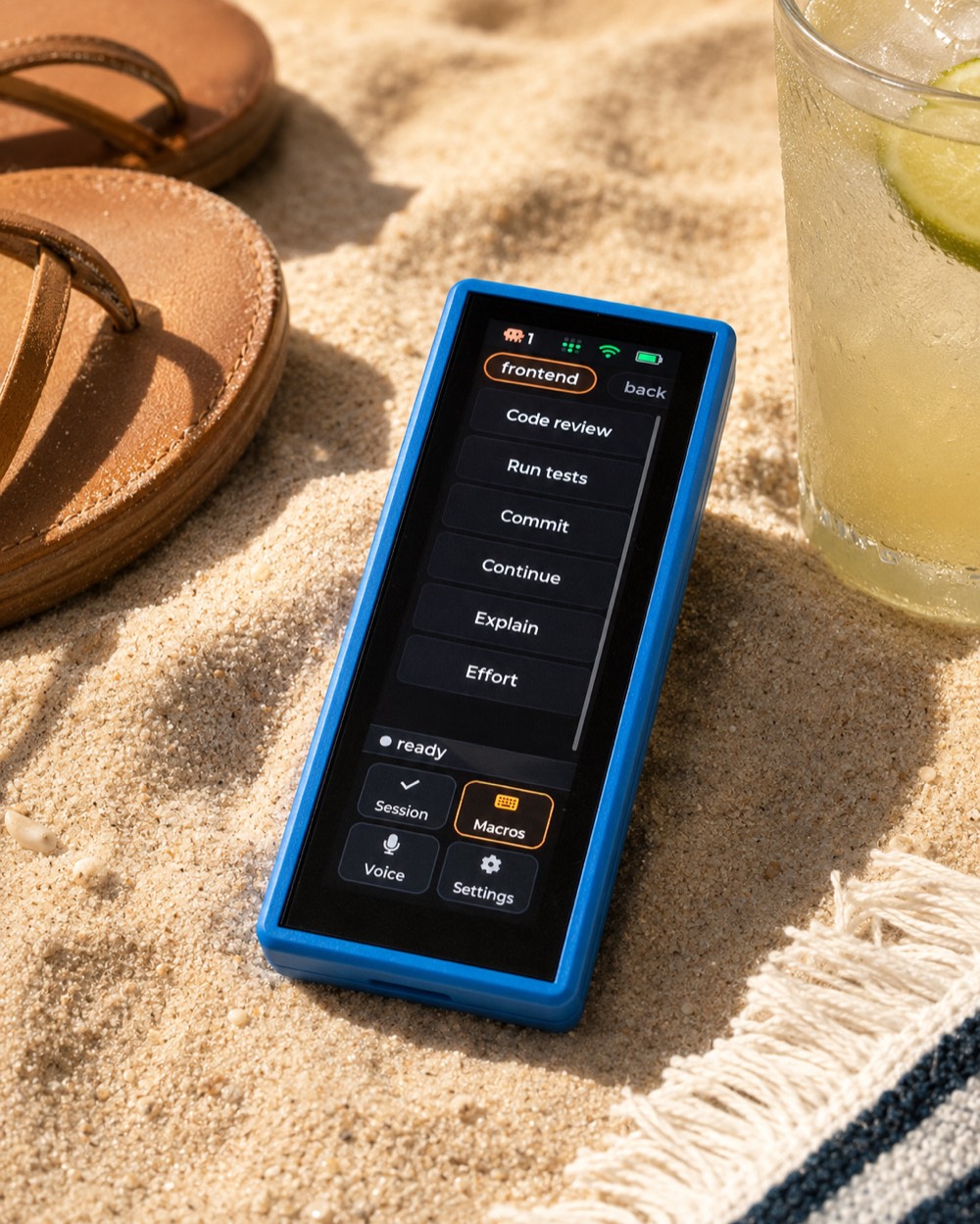 The claudeq device lying on beach sand next to sandals and a cold drink, showing the Macros deck.